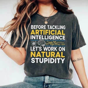 May include: Dark grey t-shirt with the text "BEFORE TACKLING ARTIFICIAL INTELLIGENCE LET'S WORK ON NATURAL STUPIDITY" in yellow and white. The shirt features a brain graphic and circuit board design.