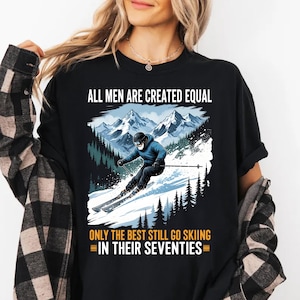 May include: Black t-shirt featuring a skier on a snowy mountain scene. The text states "ALL MEN ARE CREATED EQUAL" and "ONLY THE BEST STILL GO SKIING IN THEIR SEVENTIES". The skier is dressed in a blue jacket and black helmet.