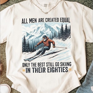 May include: Cream-colored t-shirt with a graphic of a skier on a snowy mountain slope. The skier is wearing an orange jacket and black pants. Text on the shirt reads "ALL MEN ARE CREATED EQUAL ONLY THE BEST STILL GO SKIING IN THEIR EIGHTIES."