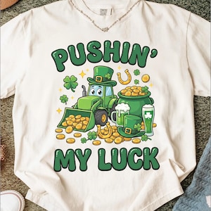 May include: White t-shirt with a St. Patrick's Day graphic. The design features a green tractor, a pot of gold, a horseshoe, and the text "PUSHIN' MY LUCK" in green. The shirt is made of a soft material and is suitable for casual wear.