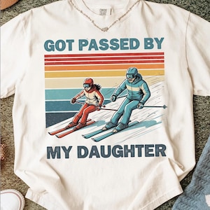 Könnte beinhalten: Cremefarbenes T-Shirt mit dem Text "GOT PASSED BY MY DAUGHTER" über einer Illustration von zwei Skifahrern. Die Skifahrer tragen rote und blaue Skianzüge vor einem Retro-Sonnenuntergangshintergrund.