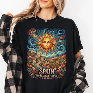 May include: Black t-shirt featuring a vibrant illustration of a sun with a face, a village, and the text "SPAIN TOTAL SOLAR ECLIPSE 8.12.2026". The design includes swirling patterns in shades of blue, orange, and green.