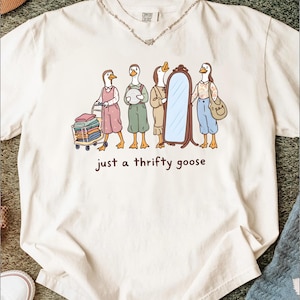 May include: Cream-colored t-shirt featuring a whimsical illustration of anthropomorphic geese. The geese are dressed in various outfits, with one pushing a shopping cart. The text "just a thrifty goose" is printed below the illustration.