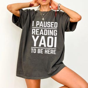 Puede incluir: Camiseta gris oscuro con el texto blanco "I PAUSED READING YAOI TO BE HERE". La camiseta tiene mangas cortas y un corte holgado.