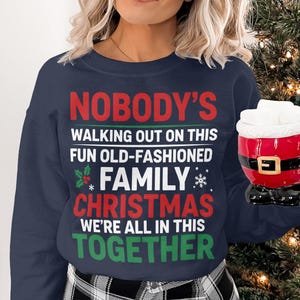 May include: Navy blue sweatshirt with the text "NOBODY'S WALKING OUT ON THIS FUN OLD-FASHIONED FAMILY CHRISTMAS WE'RE ALL IN THIS TOGETHER" in red, white, and green. A red Santa-themed mug with white foam is held in the hand.