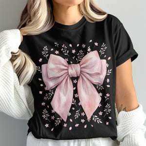 Könnte beinhalten: Schwarzes T-Shirt mit einem großen, hellrosa Schleifendesign. Die Schleife ist von kleinen weißen Blumen und grünen Blättern umgeben. Das T-Shirt hat einen klassischen Rundhalsausschnitt.