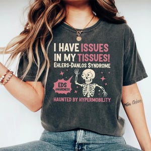 May include: A dark grey t-shirt with the text "I HAVE ISSUES IN MY TISSUES! EHLERS-DANLOS SYNDROME" in white and pink. The design includes a skeleton graphic, the words "EDS AWARENESS" and "HAUNTED BY HYPERMOBILITY".