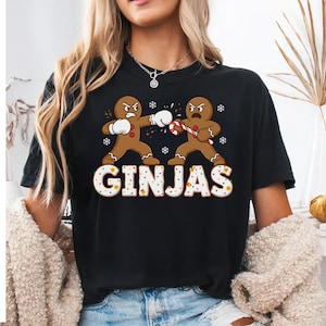 May include: Black t-shirt featuring a graphic of two gingerbread men boxing, one with a candy cane. The word "GINJAS" is displayed in a festive font. Snowflakes are scattered around the gingerbread men.