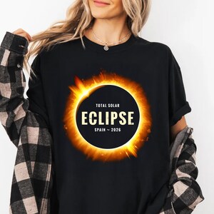 Puede incluir: Camiseta negra con un diseño de eclipse solar. El diseño incluye las palabras "TOTAL SOLAR ECLIPSE" y "ECLIPSE SPAIN ~ 2026" en un círculo, con tonos naranja y amarillo.