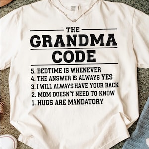 Puede incluir: Camiseta color crema con el texto "THE GRANDMA CODE" en letras negras en negrita. Debajo, se enumeran cinco reglas, incluyendo "HUGS ARE MANDATORY" y "BEDTIME IS WHENEVER". Un collar es visible en el escote.