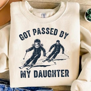 May include: A cream-coloured sweatshirt with the navy blue text "GOT PASSED BY MY DAUGHTER". The graphic shows two skiers in black and white, skiing down a snowy slope. A fun, winter-themed apparel item.
