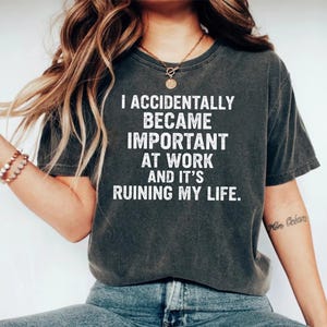 May include: Dark gray t-shirt with the white text: "I ACCIDENTALLY BECAME IMPORTANT AT WORK AND IT'S RUINING MY LIFE." The shirt is a crew neck and has short sleeves. The model is wearing blue jeans and a necklace.