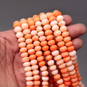May include: A collection of coral beads in shades of orange and peach. The oval-shaped beads are strung together, creating a textured and colourful display. The beads are held in a hand, showcasing their natural beauty.