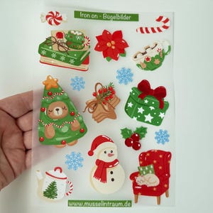 Könnte beinhalten: Ein Bogen mit weihnachtlichen Bügelbildern, darunter ein Bücherstapel, ein Weihnachtsstern, eine Tasse Kakao, ein Bär im Tannenbaumkostüm, ein Geschenk, ein Schneemann, eine Teekanne und eine Katze auf einem roten Sessel mit dem Text Iron on - Bügelbilder.