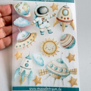 Könnte beinhalten: Ein Bogen mit Bügelbildern mit Weltraummotiven, darunter ein Astronaut, Raketen, UFOs, ein Planet, eine Sonne mit Gesicht, Wolken und Sterne. Der Text "Iron on - Bügelbilder" und "www.musselintraum.de" ist auf dem Bogen aufgedruckt.