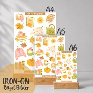 Könnte beinhalten: Drei Bögen mit Bügelbildern in den Größen A4, A5 und A6 mit herbstlichen Motiven wie Kürbissen, Sonnenblumen, Büchern, Pullovern, Teekesseln und Blättern. Der Text Iron on - Bügelbilder ist oben auf jedem Bogen aufgedruckt.