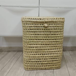 Puede incluir: Una cesta rectangular tejida con tapa, hecha de fibras de color natural. La cesta tiene una superficie texturizada y un cierre de botón. La cesta está sobre un suelo de madera de color claro. Es un contenedor de almacenamiento.