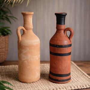 Op de afbeelding: Twee terracotta flessen met handvatten. Eén is effen, de andere is beschilderd met zwarte strepen en een zwarte bovenkant. Ze worden tentoongesteld op een geweven mat, met een houten tafel en groen in de achtergrond. De flessen zijn ongeveer 25 cm hoog.