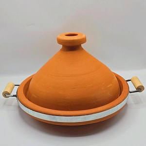 Peut inclure: Un tajine en terre cuite de couleur orange naturel, avec un couvercle conique et une base large. Une bande métallique blanche entoure la base. Des poignées en bois sont fixées sur les côtés.