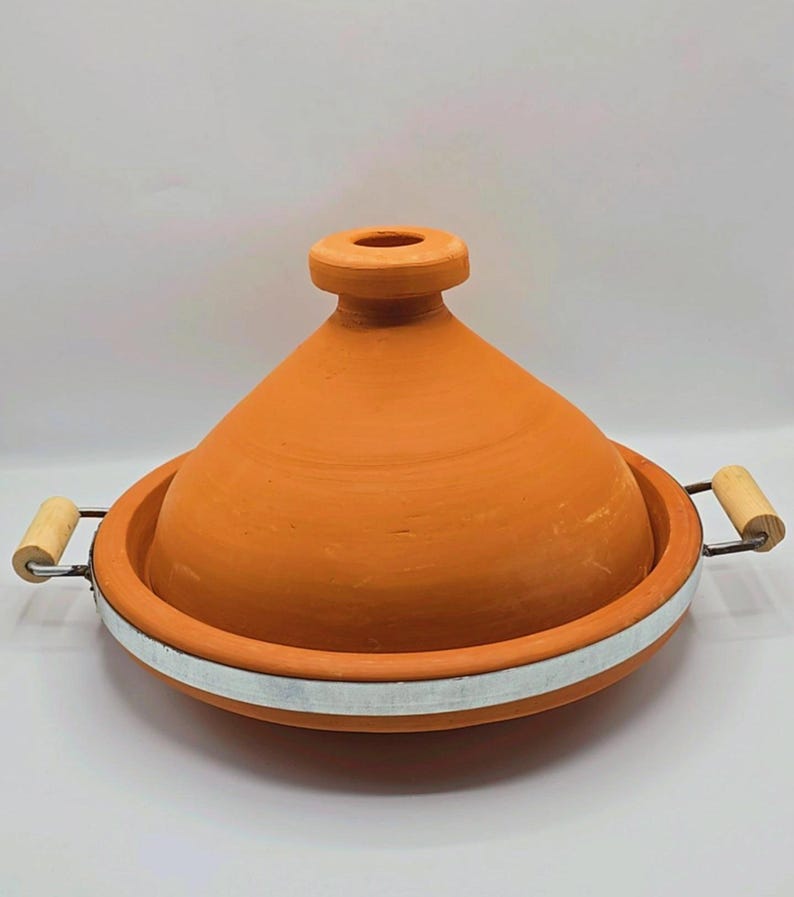 May include: A terracotta tagine pot with a conical lid and a wide, shallow base. The pot has a natural, earthy orange color, with a white metal band around the base. Wooden handles are attached to the sides.