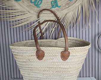 Puede incluir: Bolso tote de paja tejida con asas y detalles de cuero marrón. El bolso tiene una abertura ancha y una base resistente. Un sombrero de paja con texto turquesa está en el fondo.