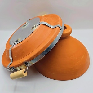 May include: An orange-brown clay cooking pot with a lid. The lid has a metal plate and a wooden handle. The pot is designed for cooking and has a round shape. The lid is partially open, revealing the interior of the pot.