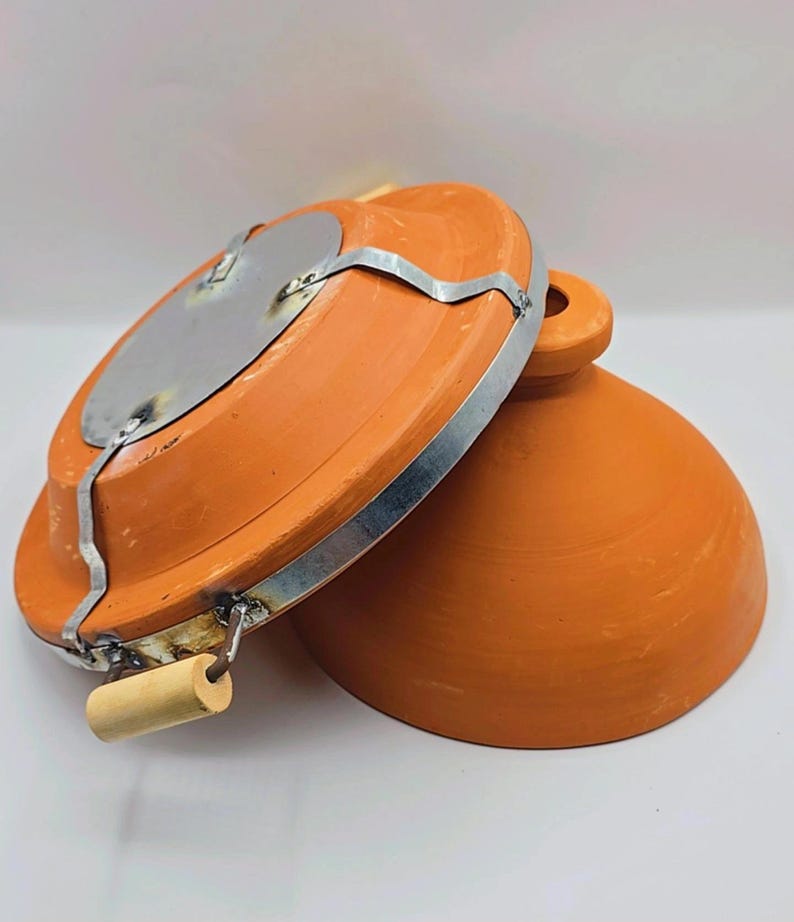 May include: An orange-brown clay cooking pot with a lid. The lid has a metal plate and a wooden handle. The pot is designed for cooking and has a round shape. The lid is partially open, revealing the interior of the pot.