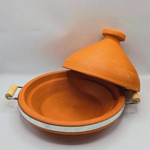May include: An open, orange-brown tagine pot with a conical lid. The earthenware pot has a wide, shallow base with a white band and wooden handles. The lid is slightly ajar, revealing the interior. A traditional cooking vessel.
