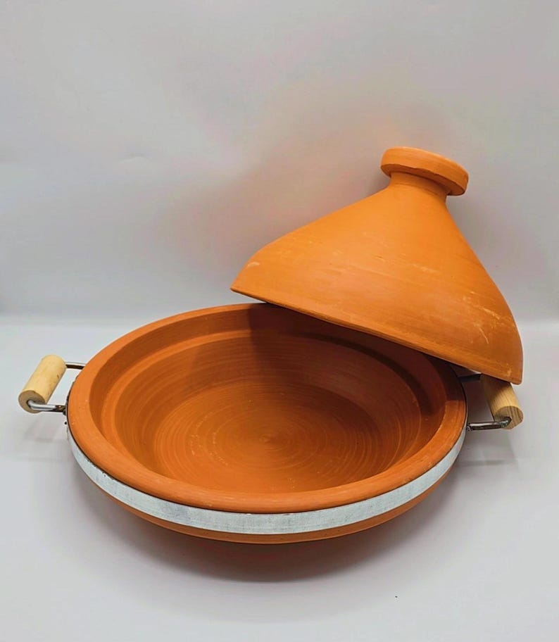 May include: An open, orange-brown tagine pot with a conical lid. The earthenware pot has a wide, shallow base with a white band and wooden handles. The lid is slightly ajar, revealing the interior. A traditional cooking vessel.