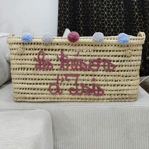 Pode incluir: Cesto retangular tecido com o texto "Les Fraises d'Isis" em fio rosa. O cesto é adornado com pompons azuis, cinzentos e rosa. O cesto é feito de material tecido de cor natural.