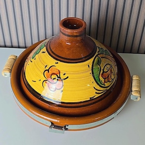 Op de afbeelding: Een keramische tajine-pot met een conische deksel en een brede, ondiepe basis. De pot is voornamelijk bruin met een gele deksel versierd met bloemmotieven in groen, oranje en zwart. Het heeft houten handvatten en een metalen sluiting.
