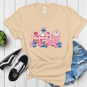 May include: Beige t-shirt featuring a graphic of cartoon characters and pink and white beverages. The design includes hearts and the text "The Love". The shirt is paired with black sneakers and denim shorts.