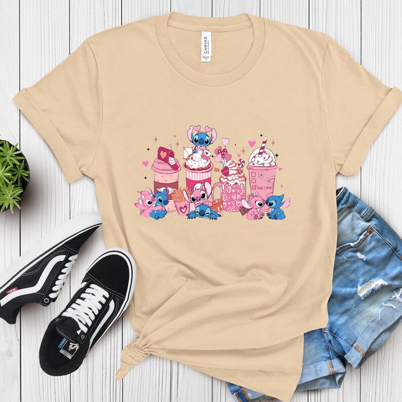May include: Beige t-shirt featuring a graphic of cartoon characters and pink and white beverages. The design includes hearts and the text "The Love". The shirt is paired with black sneakers and denim shorts.