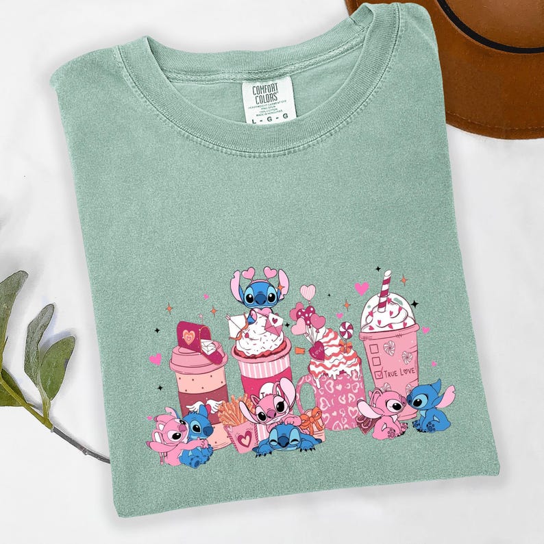 May include: A sage green t-shirt featuring a graphic of cartoon characters and pink and red desserts. The design includes pink hearts, a pink drink with a straw, and the text "True Love".