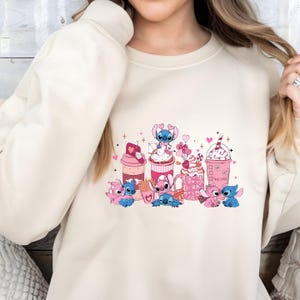 May include: Cream-colored sweatshirt featuring a colorful graphic of cartoon characters and Valentine's Day-themed treats. The design includes pink and blue characters, hearts, and various desserts like milkshakes and cupcakes. The phrase "True Love" is also visible.