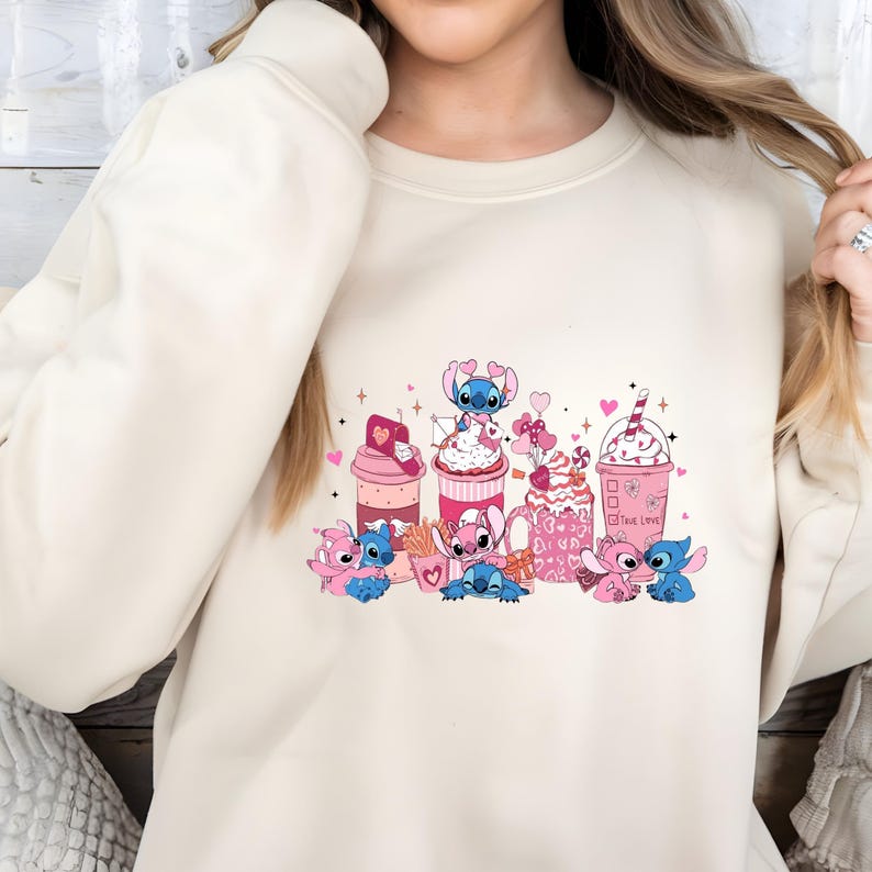May include: Cream-colored sweatshirt featuring a colorful graphic of cartoon characters and Valentine's Day-themed treats. The design includes pink and blue characters, hearts, and various desserts like milkshakes and cupcakes. The phrase "True Love" is also visible.