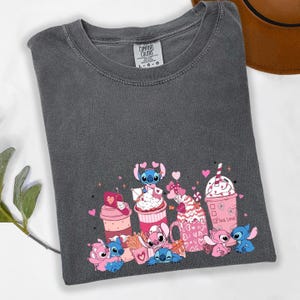 May include: Dark gray t-shirt with a graphic featuring cartoon characters, pink hearts, and various pink and white beverages. The text "True Love" is printed on one of the cups. The shirt has a Comfort Colors tag.