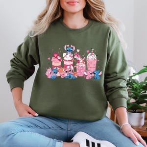 May include: An olive green sweatshirt featuring a colorful graphic of cartoon characters and desserts. The design includes pink, blue, and white elements, with hearts and the text "Tea Love". The sweatshirt is a crew neck style.