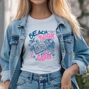 May include: White t-shirt with a blue and pink graphic that reads "Beach Book Club." The design includes seashells, a starfish, a hat, and a book. The shirt is paired with a light blue denim jacket and jeans.