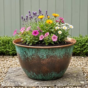 May include: A large, hammered copper planter with a verdigris finish, filled with a variety of colorful flowers, including lavender, yellow marigolds, pink petunias, and white daisies. The planter sits on a stone slab.