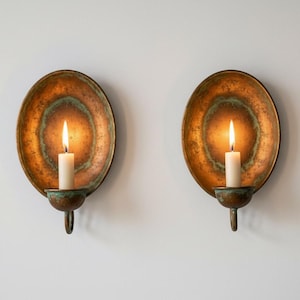 Puede incluir: Dos apliques de pared antiguos con velas encendidas. Cada aplique presenta una placa trasera ovalada de color cobre con pátina verde. Una vela blanca arde en el centro de cada aplique, proyectando un brillo cálido. Los apliques están montados en una pared blanca.