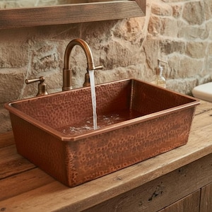 Op de afbeelding: Een rechthoekige koperen wastafel met een gehamerde textuur is gemonteerd op een houten wastafel. Een kraan giet water in de wastafel. De badkamer heeft een stenen muur en een wit toilet.