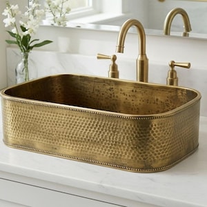 Puede incluir: Un lavabo rectangular de latón martillado con grifo y accesorios a juego. El lavabo está sobre una encimera blanca, con un mueble blanco y un espejo en el fondo. El grifo tiene un caño curvo y manijas de palanca.