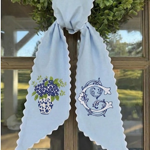 Op de afbeelding: Een lichtblauw deursjerp met een geschulpte rand en witte rand, versierd met borduursels. Eén kant toont een blauw-wit bloemstuk in een vaas, de andere een decoratieve letter "C".