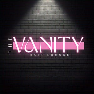 May include: A neon sign against a dark brick wall. The sign reads "THE VANITY" in white capital letters, with a pink horizontal bar behind the word. Below, in smaller white letters, it says "HAIR LOUNGE".