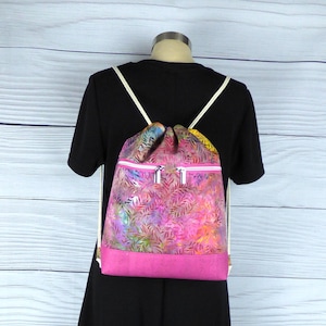 May include: A pink drawstring backpack with a floral print and two zippered pockets. The backpack is being worn by a mannequin wearing a black dress.