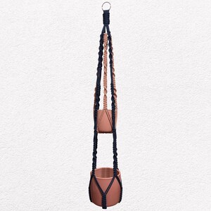 May include: A two-tiered macrame plant hanger with terracotta-colored pots. The macrame is a combination of navy blue and tan-colored rope, creating a stylish and functional piece for displaying plants. The hanger is suspended from a metal ring.