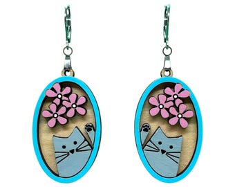 May include: A pair of wooden earrings featuring an oval design with a light blue border. Each earring showcases a gray cat with pink flowers. The earrings have silver-colored ear wires.