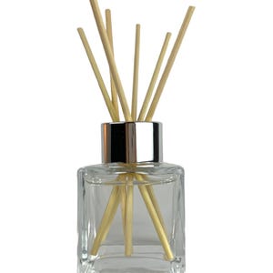 May include: A clear glass diffuser bottle with a silver collar and a bundle of light brown reeds. The square-shaped bottle is filled with a clear liquid. The reeds are arranged to fan out from the top of the bottle.