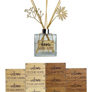 May include: A clear glass diffuser bottle with the word "home" on a gold tag, accompanied by wooden reeds and decorative accents. Below are wooden signs with "welcome" messages for various locations, including home, cabin, and campsite.